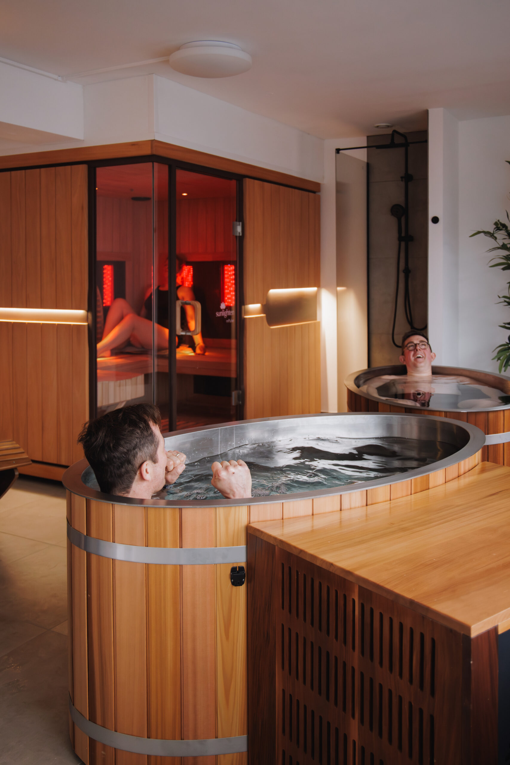 Two people relax in large wooden hot tubs, whilst another person lounges inside a modern wooden sauna in the background. The setting appears to be a spa with wood panelling and contemporary decor.
