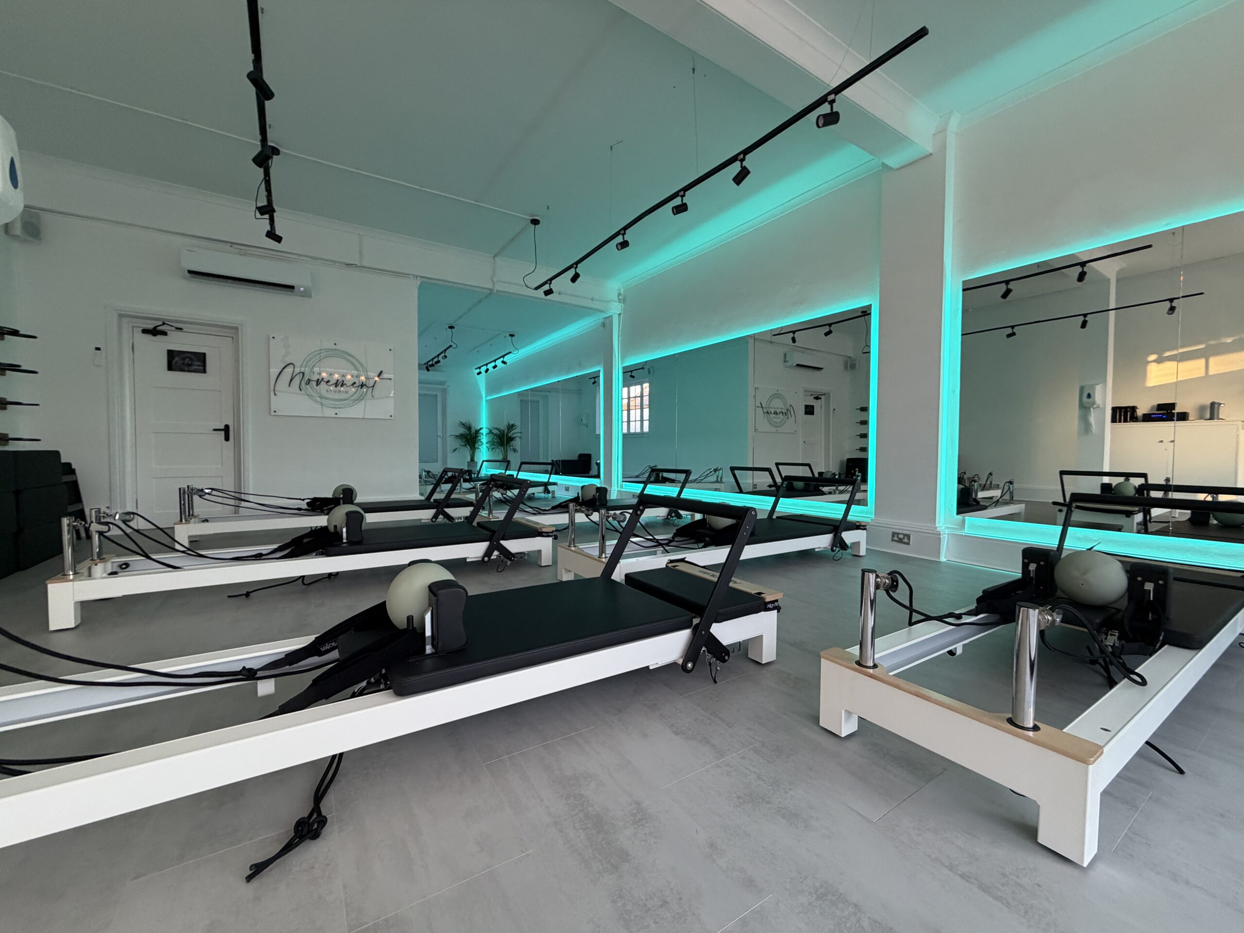 A modern Pilates studio with several reformer machines, white walls, large mirrors, and blue LED lighting creating a clean, calming atmosphere.